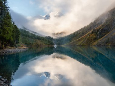 Resimli dağ sisli gölü yaz sabahında. Bulutlu gökyüzünün altındaki dağ gölünün su yüzeyinde buzul yansıması olan parlak bir manzara. Dağların yansıması, gökyüzü ve beyaz bulutlar.
