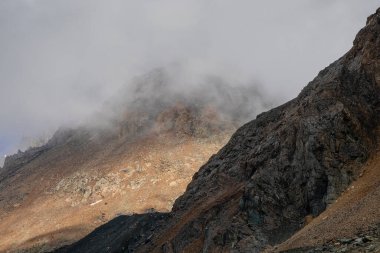 Sofr focus. Atmospheric ghostly landscape with fuzzy silhouettes of sharp rocks in low clouds. Dramatic view to large mountains blurred in rain haze in gray low clouds. Diagonal slope of the mountain.