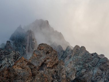 Soft focus. Atmospheric ghostly landscape with fuzzy silhouettes of sharp rocks in low clouds. Dramatic view to large mountains blurred in rain haze in gray low clouds.