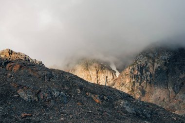 Soft focus. Atmospheric ghostly landscape with fuzzy silhouettes of golden sharp rocks in low clouds. Dramatic view to large mountains blurred in rain haze in gray low clouds. 