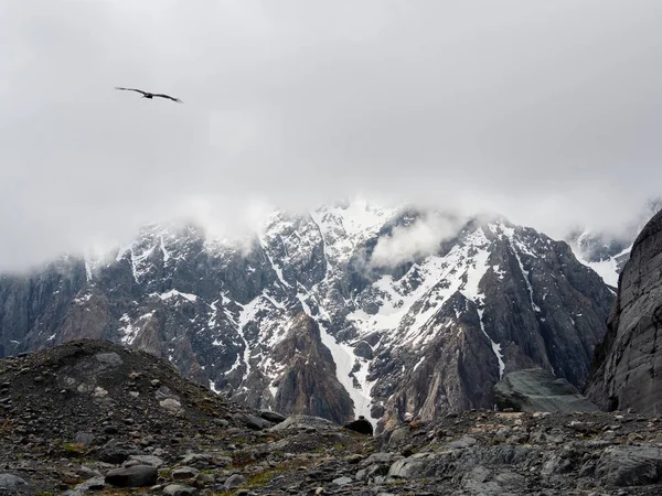 Soft focus. Atmospheric ghostly landscape with fuzzy silhouettes of snow rocks in low clouds. Dramatic view to large mountains blurred in rain haze in gray low clouds. Sharp cliff of the mountain.