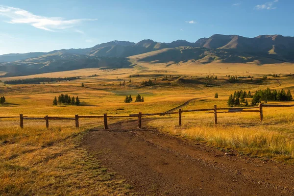 Dirt road is blocked by a fence. The path is closed, private territory. Beautiful sunny landscape with golden vast field with long fence. Vivid scenery with autumn field behind wood fence in mountains