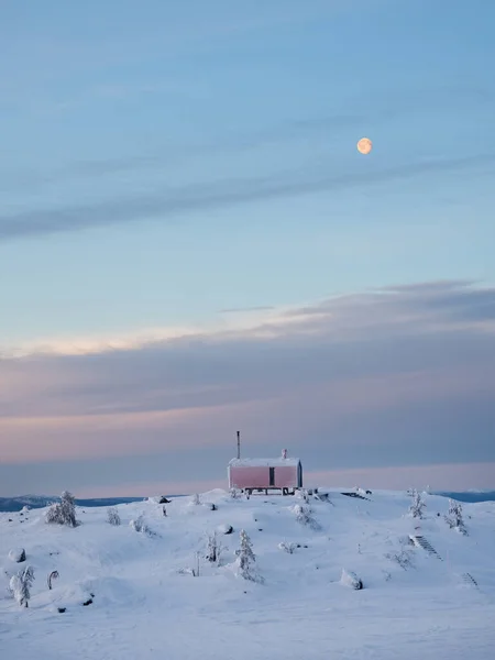 Lonely bungalow ander full moon on a snowy mountain slope under a bright dawn sky. Vertical minimalistic background with of lonely red cabin in winter.