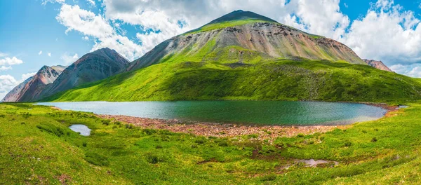 Dağlık vadide, güneş ışığında dağ gölü ve mavi bulutlu gökyüzünün altında büyük bir dağ olan açık yeşil alp manzarası. Yeşil dağ vadisinde bulutların gölgesi. Panoramik görünümler.