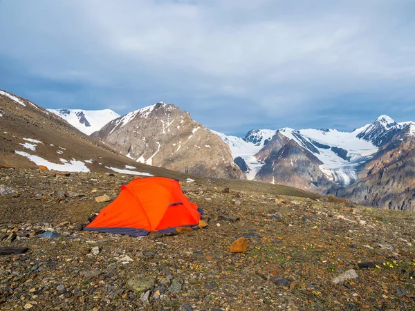 Summer camping in mountain. Bright alpine landscape with vivid orange tent at very high altitude with view to high mountain and large glacier in dark clouds. Awesome mountain scenery with tent.