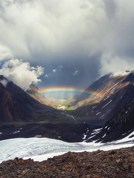 Rainbow over a mountain valley. Gloomy scenery with bright rainbow above glacier in mountain valley. Top view to colorful rainbow and low clouds in mountains in rainy and sunny weather. Vertical view.