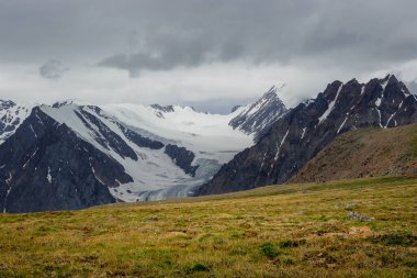 Güneşli yeşil çimen tepesinden yüksek karlı dağ sıralarına keskin tepeler ve bulutlu gökyüzünün altındaki buzullarla yaz manzarası. Değişken havada, büyük kar dağları olan renkli bir manzara..