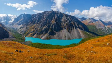 Bright alpine landscape with mountain lake in wide highland valley in sunlight and big mountain under blue cloudy sky. Shadow of clouds on autumn mountain valley. Panoramic views.