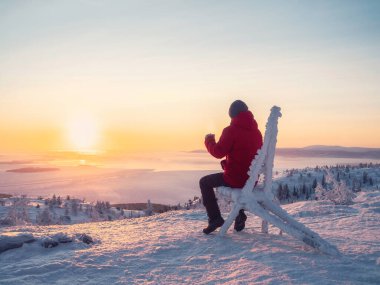 Selective focus. Happy man with a cup of tea at sunrise. View on the  frozen sea from above. Coffee from the outdoors. Snowy mountain background. Winter holidays, tourism, travel and people concept.