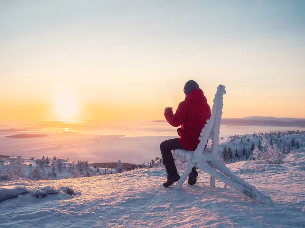 Selective focus. Happy man with a cup of tea at sunrise. View on the  frozen sea from above. Coffee from the outdoors. Snowy mountain background. Winter holidays, tourism, travel and people concept.
