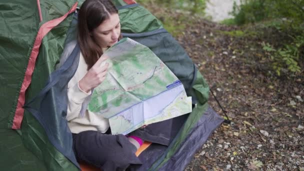 En kvinna läser en karta i ett tält i skogen. Utforska rutten för en vandring — Stockvideo
