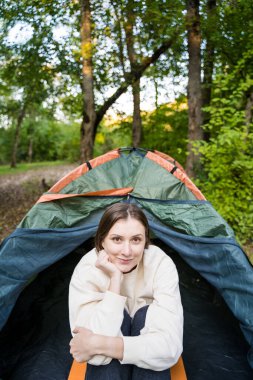 Genç turist çadırın içinde. Kadın gülümser ve kameraya bakar. Ormanda yürüyüş için rahat ve sıcak kıyafetler.