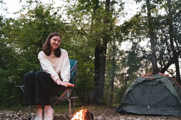 Una donna sola siede accanto al fuoco accanto a una tenda nel bosco. Riposa e si scalda durante un'escursione. Crepuscolo in natura — Foto Stock