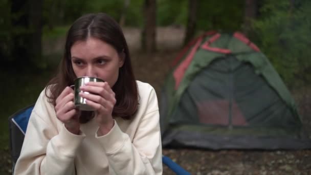 Μια νεαρή γυναίκα πίνει ένα ζεστό ποτό από μια κούπα σε μια κατασκήνωση. Άνεση στην πεζοπορία. Ζέσταμα σε κρύο έξω — Αρχείο Βίντεο