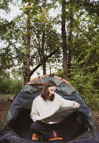 Uma turista feminina estuda a marcha em um mapa enquanto está sentada em uma tenda na floresta Fotografia De Stock
