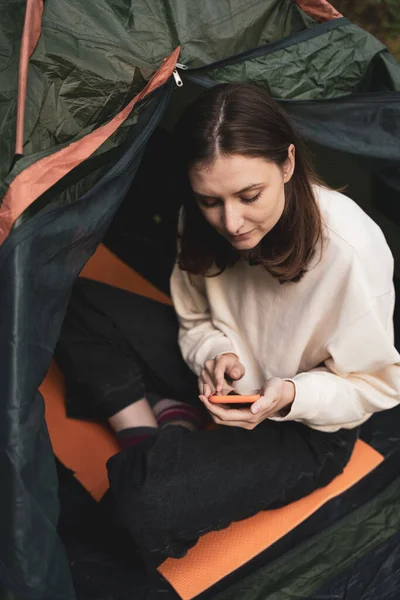 Um jovem turista senta-se em uma tenda na natureza e olha para o telefone. Navegar na Internet fora da cidade em caminhada Imagem De Stock