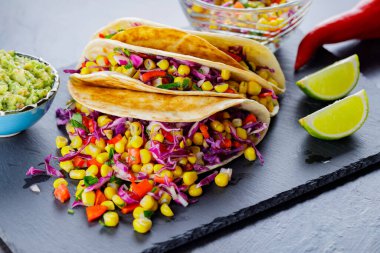 Çeşitli sebzelerle vejetaryen tacoları, guacamole ve koyu arkaplanda dilimlenmiş limon. Tatlı mısırlı taco, mor lahana ve karabiber tahtada. Boşluğu kopyala
