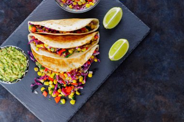 Çeşitli sebzelerle vejetaryen tacoları, guacamole ve koyu arkaplanda dilimlenmiş limon. Tatlı mısırlı taco, mor lahana ve karabiber tahtada. Boşluğu kopyala