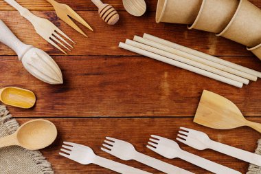Ahşap masada farklı ahşap mutfak malzemeleri. Tahta çatal bıçaklar eski tahtalarda. Bambu pipetleri ve tek kullanımlık kağıt bardaklar. Çevre dostu sofra takımı. Sıfır atık. Üst görünüm
