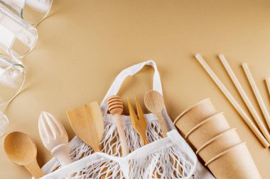 Ahşap çatal bıçak takımı ve bej arka planda ip torbası. Dünya renk arka planında tek kullanımlık kağıt bardaklar ve cam eşyalar. Sıfır atık. Uzayı kopyala Üst görünüm