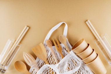 Ahşap çatal bıçak takımı ve bej arka planda ip torbası. Dünya renk arka planında tek kullanımlık kağıt bardaklar ve cam eşyalar. Sıfır atık. Uzayı kopyala Üst görünüm