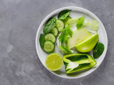 Gri arka planda dilimlenmiş yeşil meyve ve sebzeler. Beyaz tabakta taze yeşil diyet yemeği. Sağlıklı vejetaryen gıda konsepti. Uzayı kopyala Üst görünüm