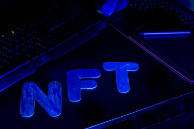 Mavi neon ışıkla aydınlatılmış harflerin NFT (Parasız Token) metni. NFT dijital sanat yaratan bir grafik tasarımcısının işyeri. Kriptoman kavramı