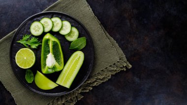 Koyu bir arka planda yeşil sebzeler ve meyveler. Çarşaf peçetesinde diyet yemeği. Siyah tabakta sağlıklı vejetaryen yemeği. Üst görünüm
