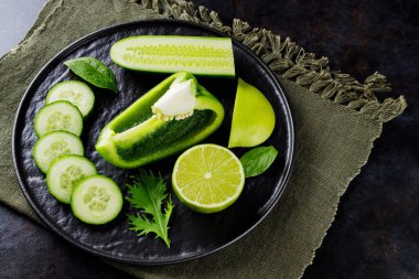 Koyu bir arka planda yeşil sebzeler ve meyveler. Çarşaf peçetesinde diyet yemeği. Siyah tabakta sağlıklı vejetaryen yemeği. Üst görünüm