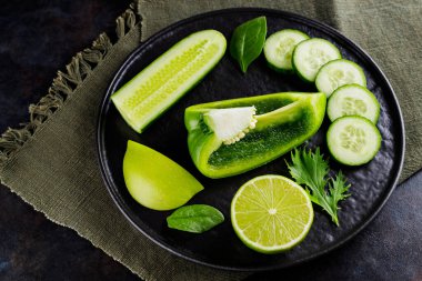 Koyu bir arka planda yeşil sebzeler ve meyveler. Çarşaf peçetesinde diyet yemeği. Siyah tabakta sağlıklı vejetaryen yemeği. Üst görünüm