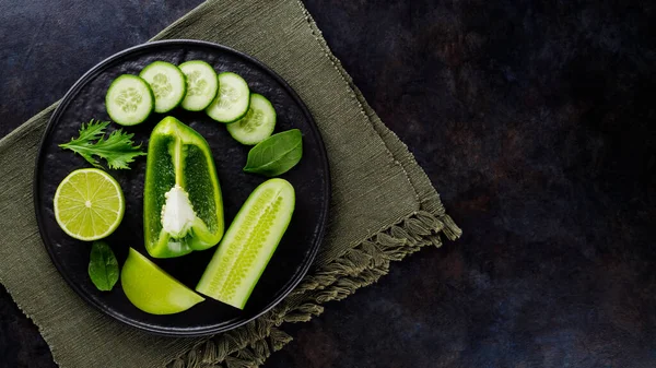 Koyu bir arka planda yeşil sebzeler ve meyveler. Çarşaf peçetesinde diyet yemeği. Siyah tabakta sağlıklı vejetaryen yemeği. Üst görünüm