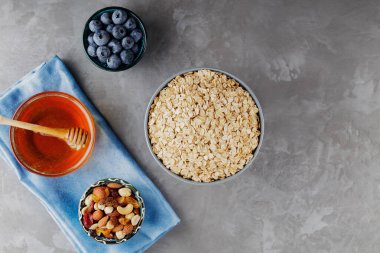 Böğürtlenli, fındıklı ve ballı sağlıklı kahvaltı. Yulaf gevreği, yaban mersini, bal ve gri arka planda çeşitli fındıklar. Yulaf ezmeli vejetaryen kahvaltısı. Üst görünüm