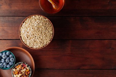 Tahta tahtalar üzerinde kahvaltı için çeşitli lezzetli malzemeler. Kuru yulaf ezmesi, yabanmersini, bal ve ahşap arka planda çeşitli fındıklar. Uzayı kopyala Üst görünüm