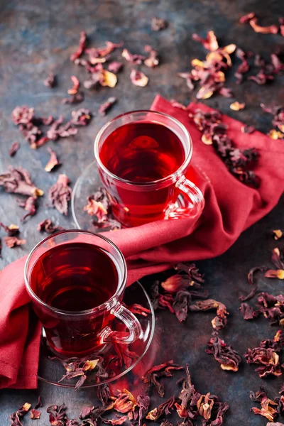Hibiscus tea in glass cup and a burgundy napkin. Cup of hibiscus tea and dry hibiscus petals on a dark background. Healthy natural slimming drink