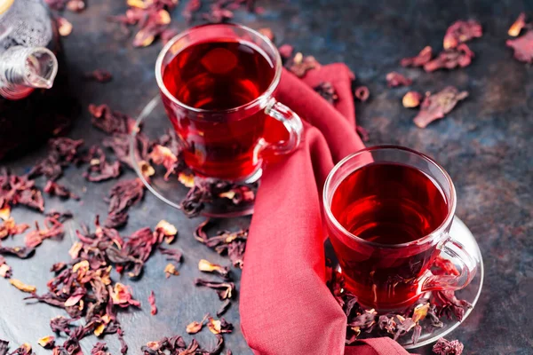 Hibiscus tea in glass cup and a burgundy napkin. Cup of hibiscus tea and dry hibiscus petals on a dark background. Healthy natural slimming drink