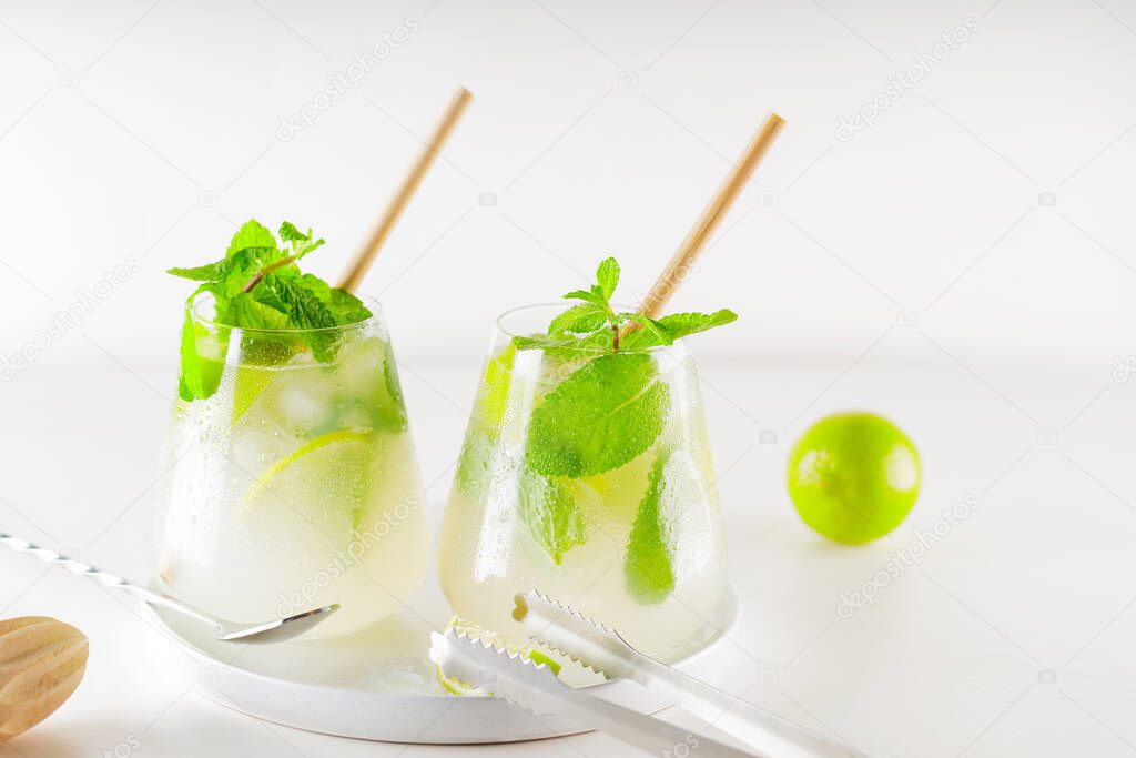 Mojito cocktail with lime and mint on white background. Two glasses of mojito with straws. Summer refreshing cocktail and bar accessories on a white plate. Copy space