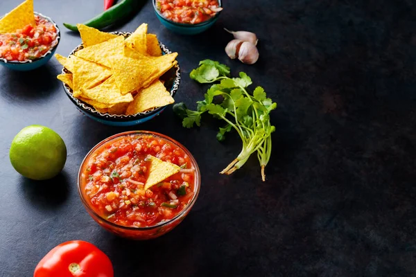 Salsa sauce and ingredients: tomato, onion, garlic, cilantro, lime, pepper. Homemade mexican salsa sauce and nacho chips on a dark background. Top view. Copy space