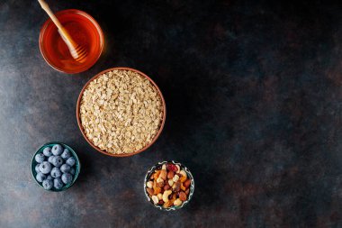 Koyu arkaplanda yulaf ezmesi, fındık, böğürtlen ve bal var. Yulaf ezmesi ve yaban mersinli sağlıklı kahvaltı için gerekli malzemeler. Uzayı kopyala Üst görünüm