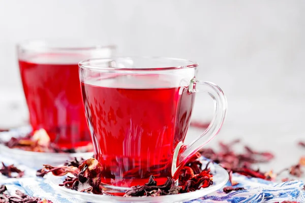 Two glass cups of hibiscus tea on a blue napkin. Hibiscus tea and dry hibiscus petals on a gray background. Healthy trending drink concept