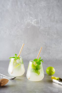 Mojito kokteyli ve gri arka planda bar aksesuarları. İki bardak mojito, kokteyl kaşığı ve buz tong. Limonlu ve naneli kokteyl. Boşluğu kopyala