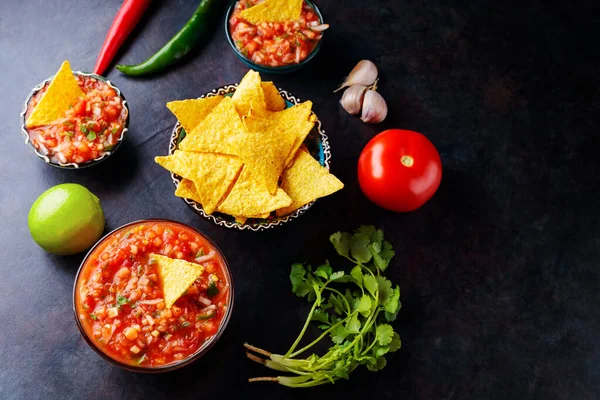 Salsa sauce and ingredients: tomato, onion, garlic, cilantro, lime, pepper. Homemade mexican salsa sauce and nacho chips on a dark background. Top view. Copy space