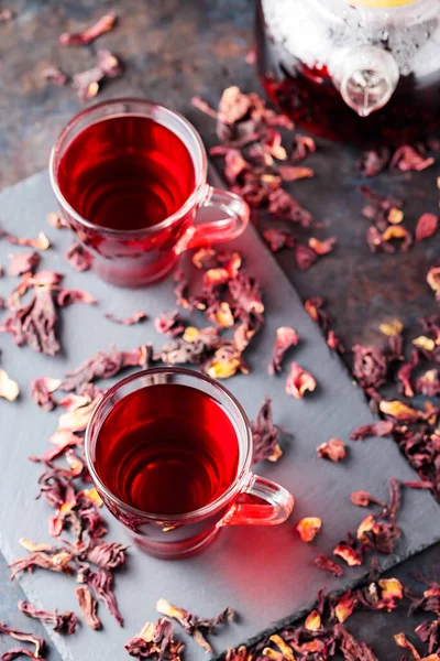 Hibiscus tea in glass mug on a dark background. Cup of red hibiscus tea and dry hibiscus petals on a slate board