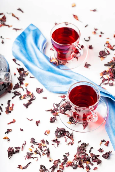 Hibiscus tea in glass cup on a blue napkin. Cup of red hibiscus tea and dry hibiscus petals on a white background. Natural dietary herbal tea