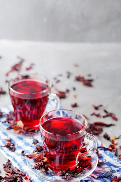 Hibiscus tea and dry hibiscus petals on gray background. Two glass cups of red hibiscus tea on blue napkin. Top view. Copy space