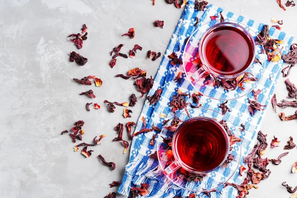 Hibiscus tea and dry hibiscus petals on gray background. Two glass cups of red hibiscus tea on blue napkin. Top view. Copy space