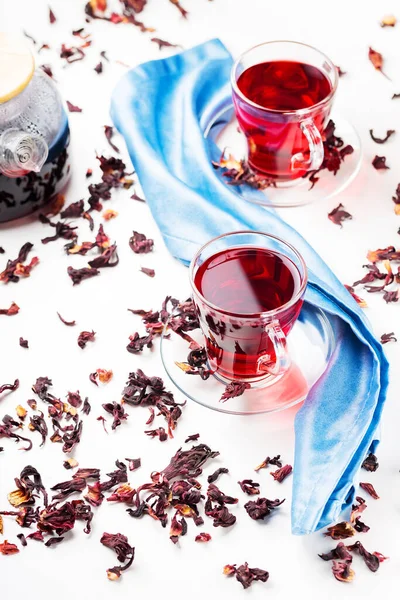 Hibiscus tea in glass cup on a blue napkin. Cup of red hibiscus tea and dry hibiscus petals on a white background. Natural dietary herbal tea