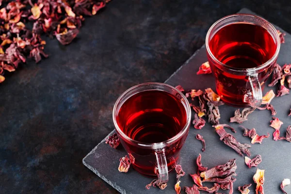 Red hibiscus tea in glass mug on a dark background. Cup of hibiscus tea and dry hibiscus petals on a slate board. Copy space