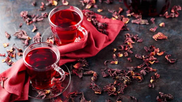 Hibiscus tea in glass cup and burgundy napkin. Cup of red hibiscus tea and dry hibiscus petals on dark background. Healthy slimming drink. Copy space