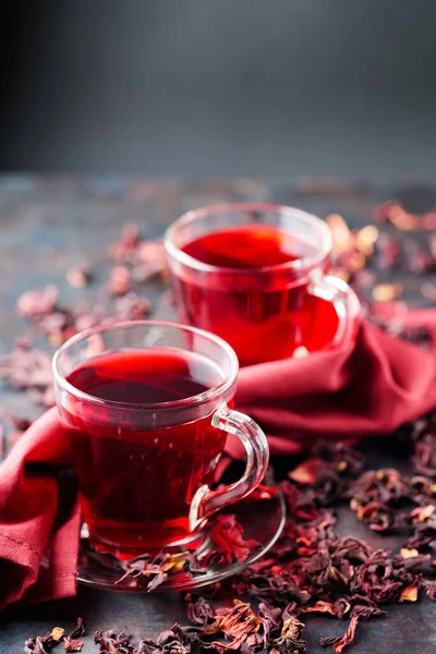 Hibiscus tea in glass cup and burgundy napkin. Cup of red hibiscus tea and dry hibiscus petals on dark background. Healthy slimming drink. Copy space