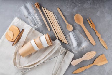 Ahşap çatal bıçak takımı, kağıt bardaklar ve gri arka planda keten çanta. Naylon poşet, bambu pipet ve ahşap sofra takımı. Geri dönüşüm veya çevre dostu kavram